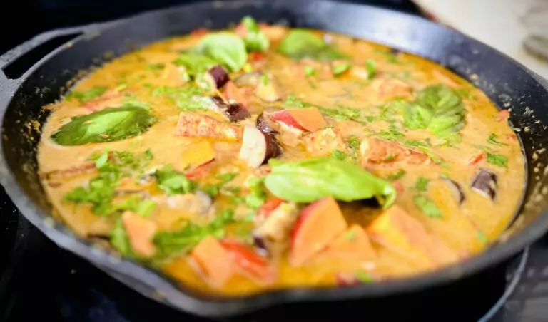 Vegan Thai green curry simmering in a cast iron skillet with sweet potato, eggplant, tofu, and fresh basil in coconut milk