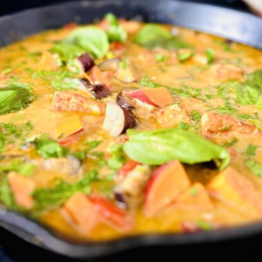 A vibrant Thai Green Curry served in a rustic cast iron pan. The curry features a creamy green-hued coconut milk base filled with cubes of golden-brown tofu, orange sweet potato chunks, purple eggplant pieces, and bright red bell pepper slices. Fresh Thai basil leaves float on top with a sprinkle of lime wedges on the side, creating a colorful, aromatic dish that showcases both the heartiness and freshness of homemade vegan Thai cuisine.