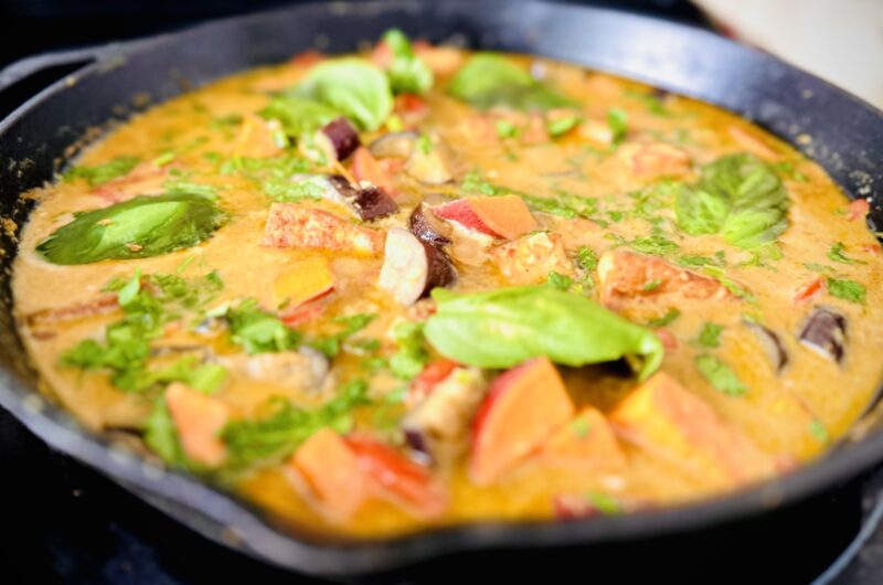 A vibrant Thai Green Curry served in a rustic cast iron pan. The curry features a creamy green-hued coconut milk base filled with cubes of golden-brown tofu, orange sweet potato chunks, purple eggplant pieces, and bright red bell pepper slices. Fresh Thai basil leaves float on top with a sprinkle of lime wedges on the side, creating a colorful, aromatic dish that showcases both the heartiness and freshness of homemade vegan Thai cuisine.
